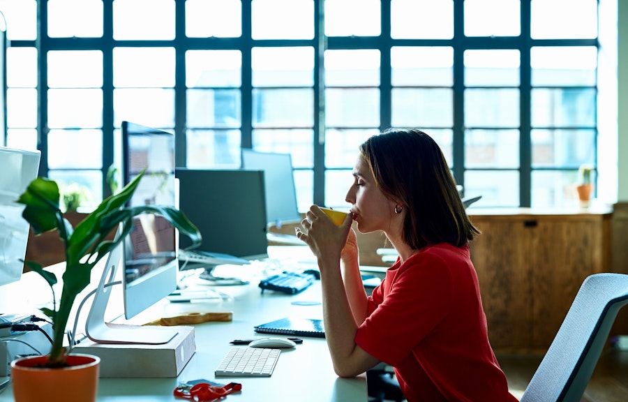 A calm looking woman in a bright office space takes a sip of her drink, indicating stepping back and control (or self-regulating) her emotions. 
