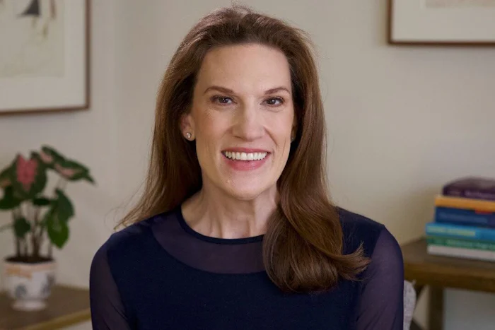 A head and shoulders portrait of Morra Aarons-Mele, a white woman with long brown hair, in an office with a plant, books and pictures.