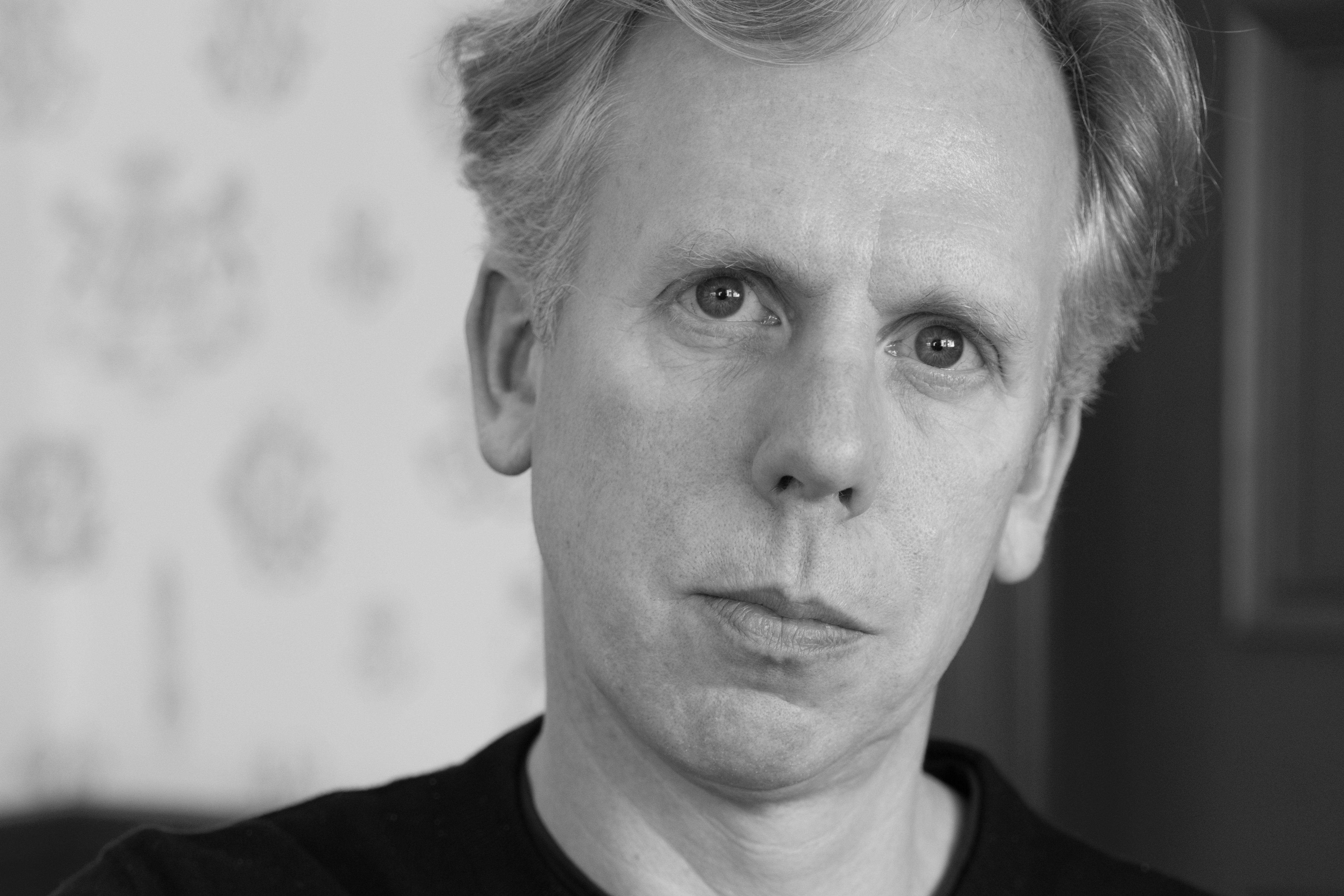 A black-and-white portrait of Alex Hill, a white man with light hair, tilting his head to one side.