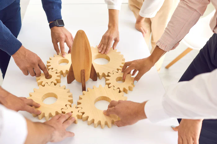 Team of cropped company employees join gearwheels around toy rocket as metaphor for unity, teamwork, choosing new effective strategy and launching new project.