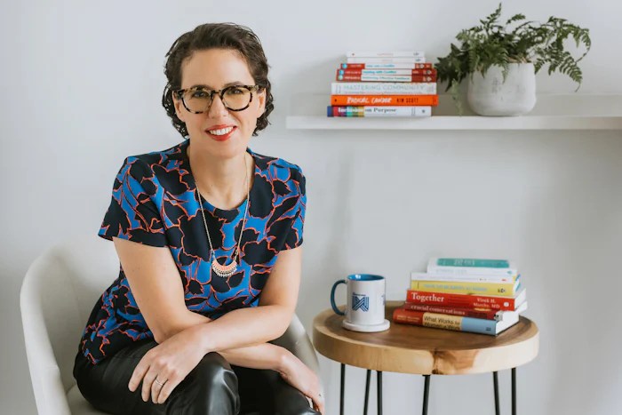 Amy Gallo, a white woman with glasses and short brown hair, sits beside a table with books and a mug, beside a shelf with a plant and more books.