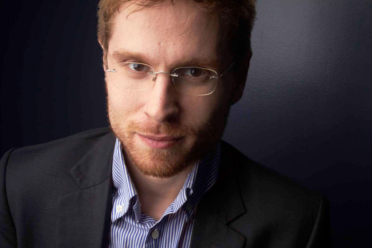 A head and shoulder portrait of Liam Martin, a white man with glasses, wearing a collared shirt and blazer.