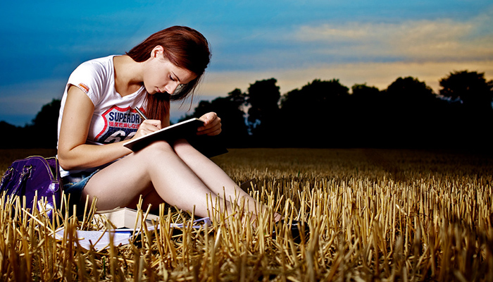 A girl sitting in a field writing in her notebook.